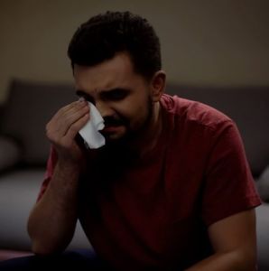 Overemotional young man watching soap opera at home and crying, wiping tears