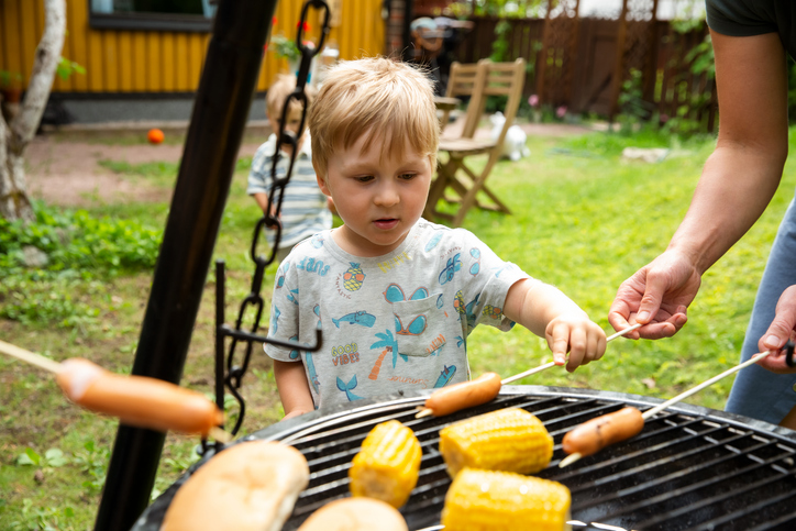 Mother grilling hot dogs and corn with toddlers on a charcoal grill. Cozy, real family moment with homemade food and warm atmosphere.