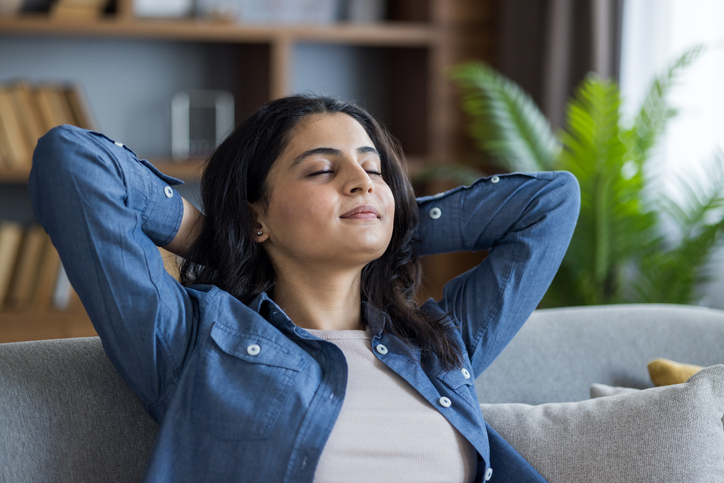 Close-up photo of a young Indian woman sitting on the couch at home, holding her hands behind her head, eyes closed, resting and napping