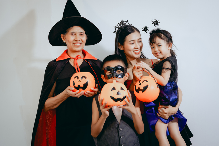 An Asian elderly woman dressed as a witch, a young Asian boy wearing a black mask, and an Asian mother with her adorable little daughter holding an orange pumpkin celebrate Halloween together